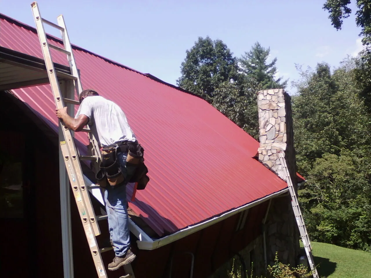 Roofer on a ladder beside a red metal roof and stone chimney — Soffit & Fascia Repair by Roofing Brothers in Dorchester